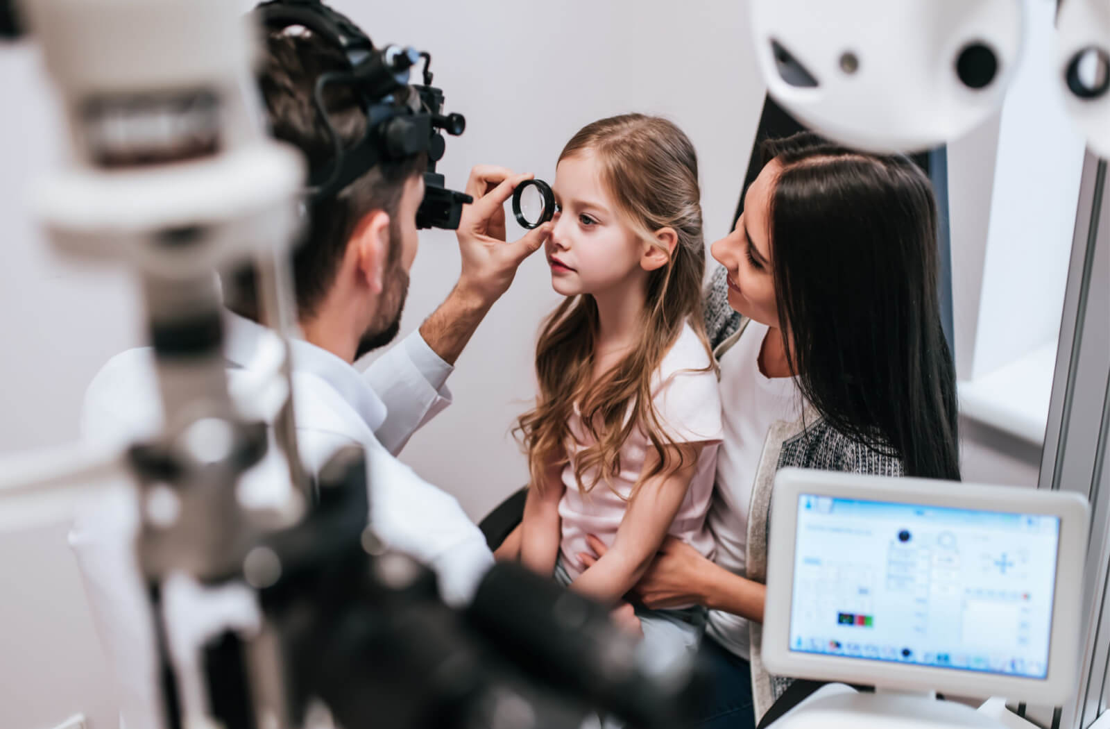 a child sits on their mother's lap while an optometrist has her look through a lens for vision therapy