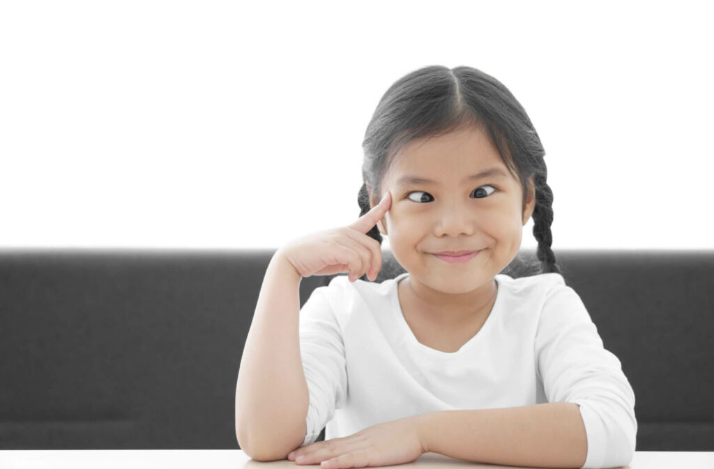 A little girl in a white shirt whit her left hand on the table while her right-hand index finger is pointing to her crossed eyes.