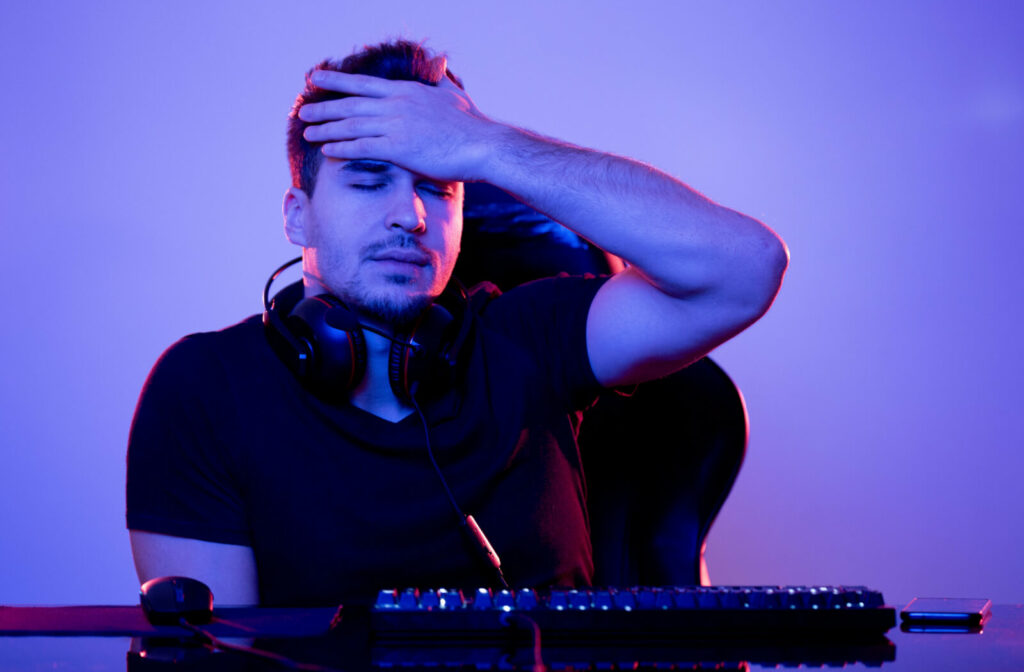 An adult male gamer sitting in front of his gaming computer and touching his forehead while his eyes are closed.