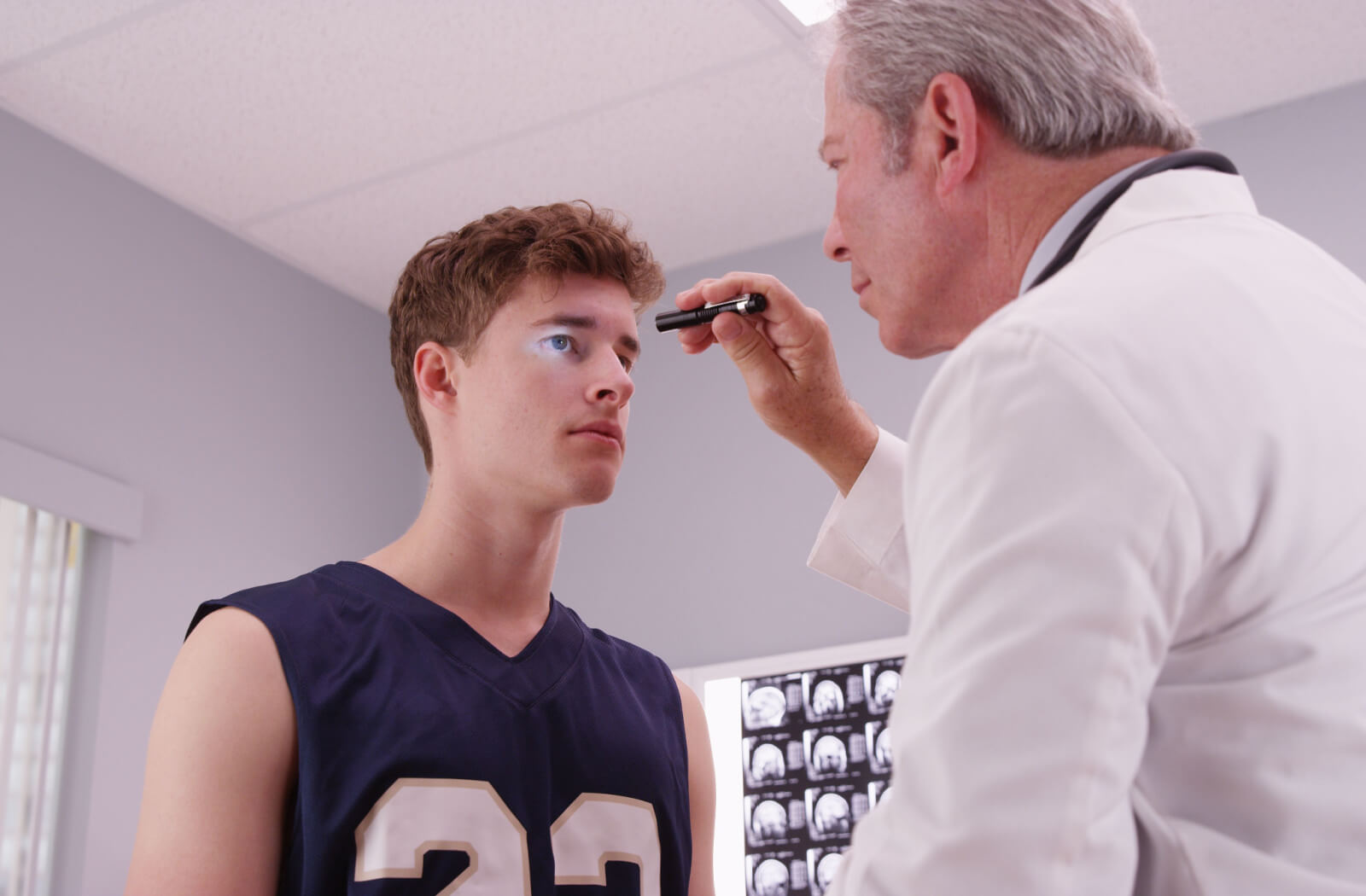 A male senior doctor is checking a basketball player's eyes with a flashlight in the clinic after a concussion during play.