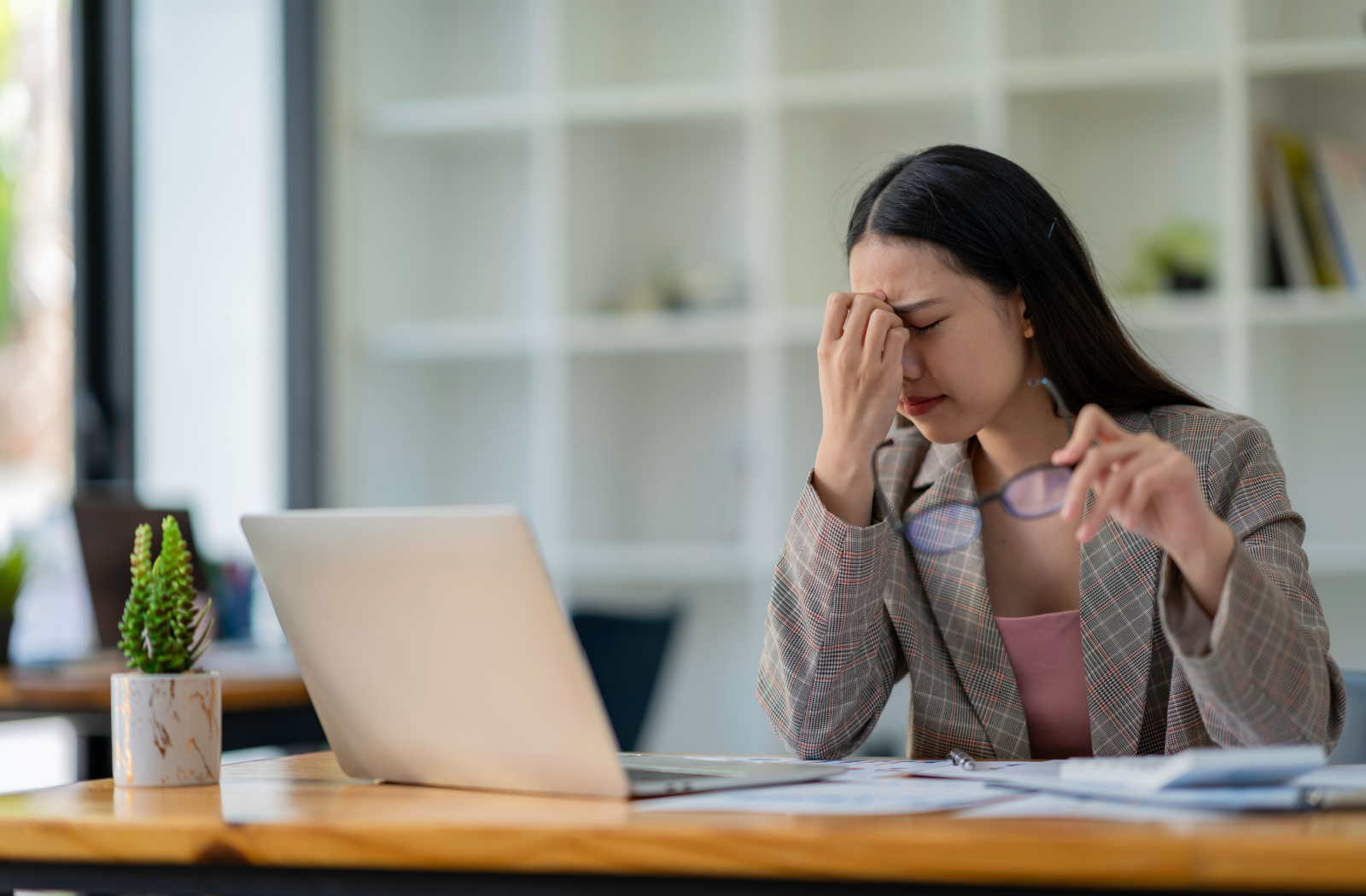 A woman in an office holding her glasses and rubbing her eyes because of dry eye symptoms related to the work she's doing on a laptop.