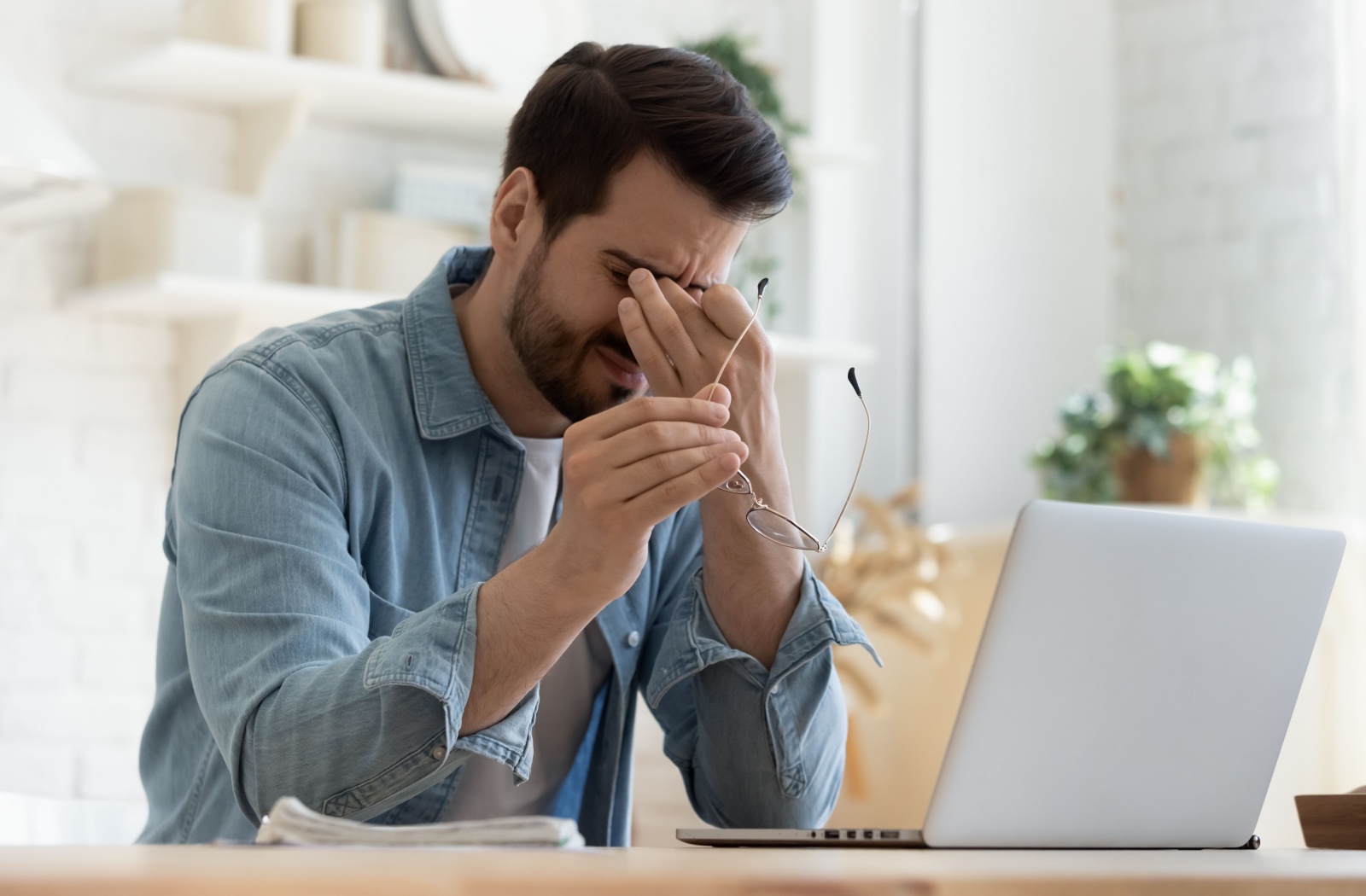 A young man sitting at a desk with his laptop, squeezing the bridge of his nose as he squeezes his eyes shut and holds his glasses in his right hand