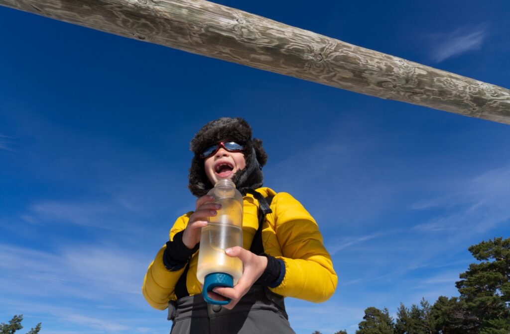 A laughing child in yellow snow gear wearing protective sunglasses while holding a large water bottle.