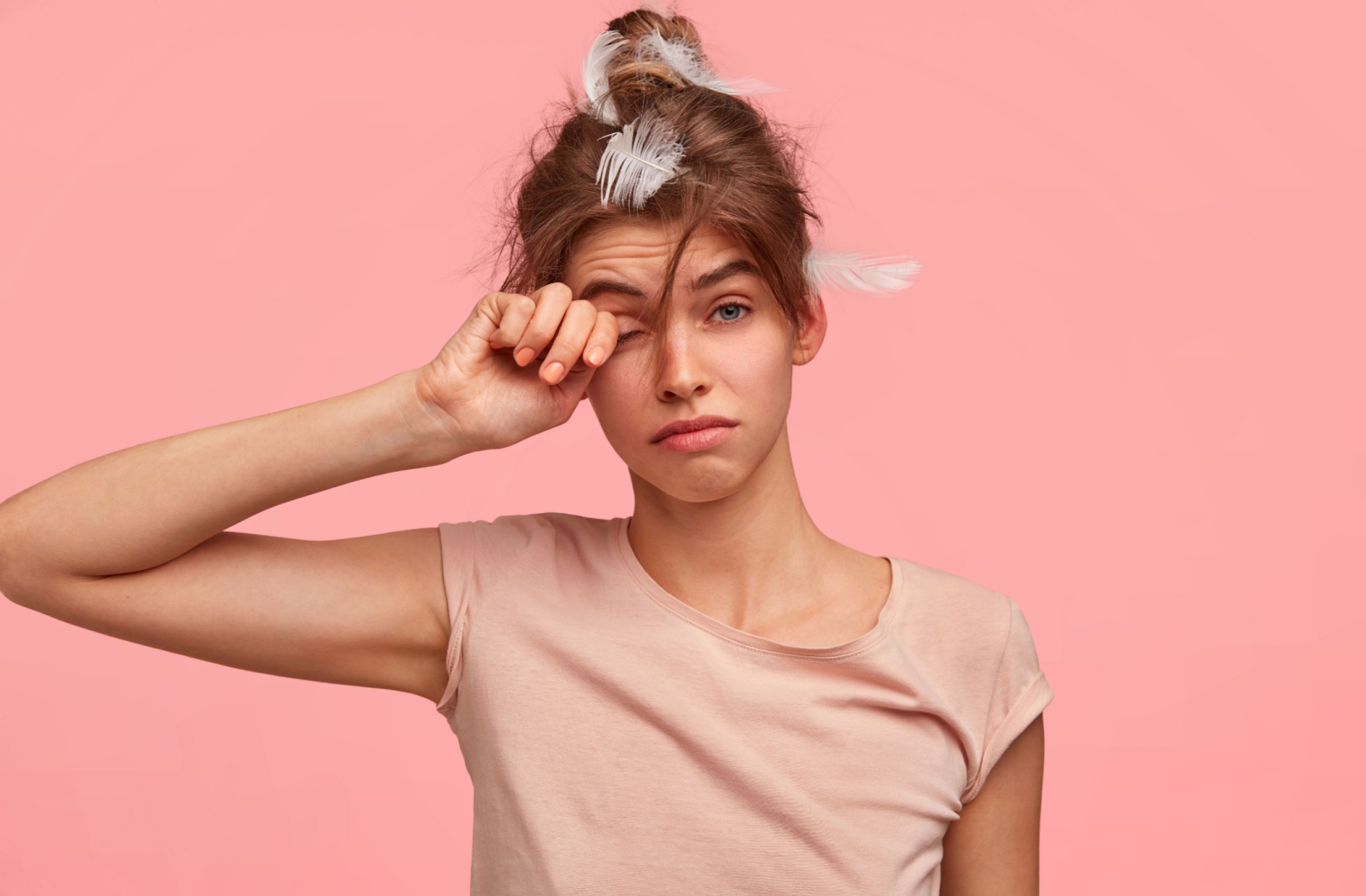 A young person with pillow feathers in their hair, rubs their eye after waking up from a sudden sleep while wearing their contact lenses.