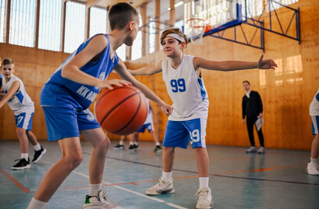 Children play basketball in a gym while one player dribbles and another defends.