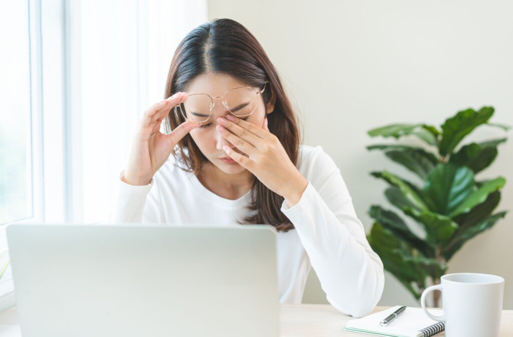 Woman sitting at a desk with her laptop, holding her glasses and rubbing her eyes, showing signs of digital eye strain or fatigue.
