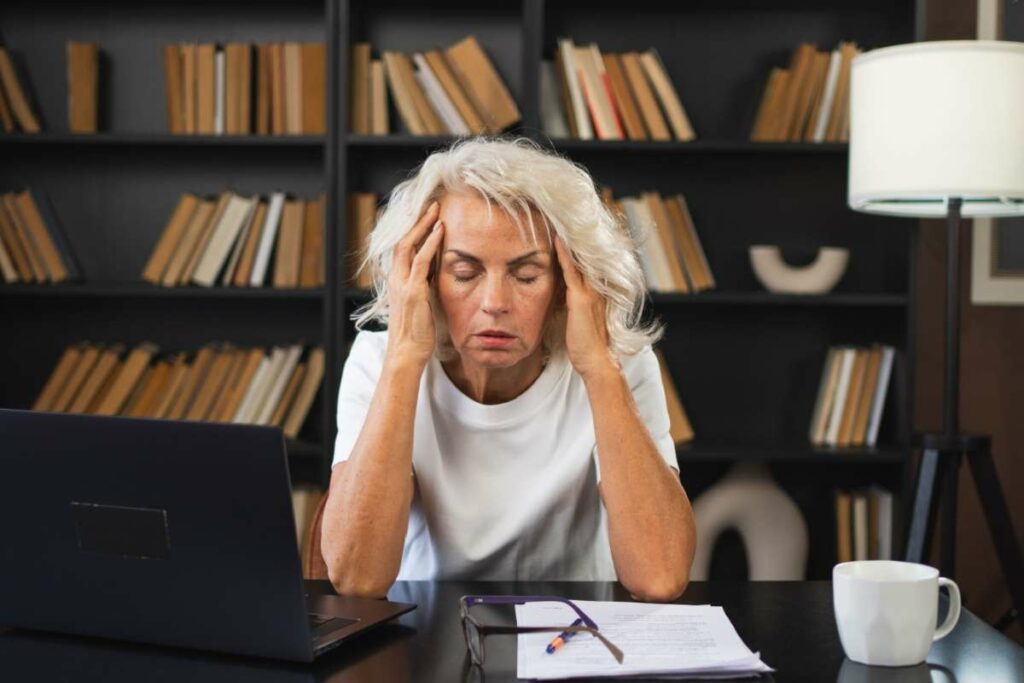 Mature woman in white shirt holding head with both hands showing signs of tension headache while sitting at desk with laptop, papers, and eyeglasses in home office with bookshelves.