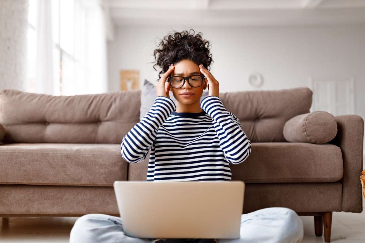 Woman in striped shirt sitting on floor holding temples with both hands showing headache symptoms while working on laptop in front of beige couch.